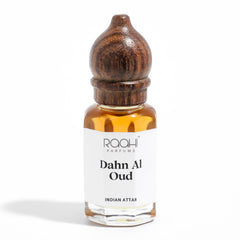 A small clear glass bottle with a dark brown wooden cap contains amber indian dahn al oud attar by raqhi parfums handcrafted in kannauj