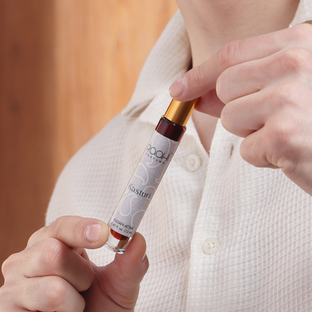 A sleek transparent glass bottle with a gold cap and maroon seal containing roohi parfums kasturi indian attar 12ml