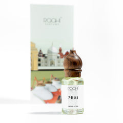 A clear glass bottle with a dark brown wooden stopper labeled mitti indian attar by roohi parfums handcrafted in kannauj