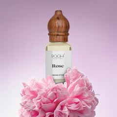 A clear glass bottle of roahi parfums rose indian attar with a wooden cap and pink peonies surrounding it